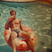 Décoration murale Wandkraft : Homme sur un matelas gonflable fleuri avec un verre à facettes, détendu dans une piscine ensoleillée. Ambiance estivale animée, décoration murale idéale. pour un intérieur élégant