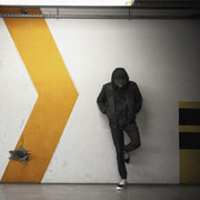 Décoration murale Wandkraft : Fresque murale urbaine avec silhouette en vêtements sombres et capuche sur fond de mur à grande flèche jaune, style urbain moderne. pour un intérieur élégant